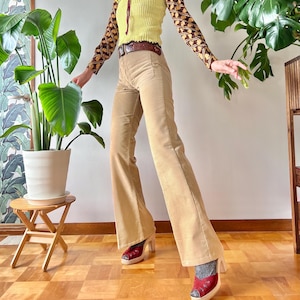 Puede incluir: Conjunto femenino que incluye un chaleco de punto amarillo con ribetes marrones, combinado con una camisa de manga larga estampada, pantalones de pana y botas rojas de tacón. Un cinturón marrón completa el look. El fondo presenta plantas de interior.