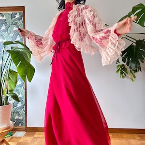 Könnte beinhalten: Ein leuchtend rotes Maxikleid mit hohem Ausschnitt und passendem Gürtel. Ein zartes, hellrosa Spitzenbolero mit Rüschenärmeln wird über dem Kleid getragen. Das Kleid ist aus einem fließenden Stoff gefertigt.
