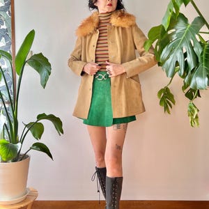 May include: A tan suede coat with a faux fur collar, paired with a green mini skirt and black lace-up boots. The outfit includes a striped turtleneck and a gold-tone belt. The ensemble is set against a neutral background with potted plants.