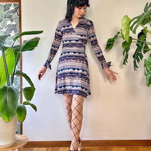 Puede incluir: Un vestido cruzado de estilo vintage con mangas largas y un estampado geométrico en tonos azules, rosas y beige. El vestido presenta un escote en V y puños con volantes. La modelo lleva medias de rejilla negras y sandalias burdeos con plataforma.