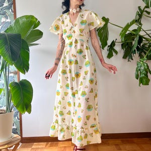 May include: A vintage-style, cream-colored maxi dress with a whimsical print of colorful landscapes. The dress features a V-neck, flutter sleeves, and a ruffled hem. The model is wearing burgundy sandals. The dress is made of a lightweight fabric.