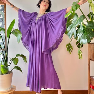 May include: A flowing, floor-length purple dress with wide, draped sleeves and a delicate lace trim. The dress is paired with burgundy sandals with a white heel. The model is wearing a silver necklace.