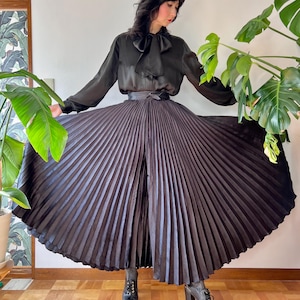 May include: A black pleated skirt with a matching black blouse featuring a bow tie neckline. The skirt is full and flowing, and the outfit is paired with black boots. The outfit is a vintage style.