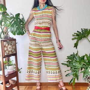 Puede incluir: Un mono blanco con un estampado geométrico de colores. El mono tiene un corpiño sin mangas, una cremallera y piernas anchas. La modelo lleva un cinturón rojo y sandalias marrones.
