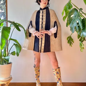 May include: A tan cape with navy blue vertical stripes and decorative cutouts. The cape is worn over a white blouse with red trim. The outfit is paired with floral print knee-high boots with blue accents. The overall style is vintage.