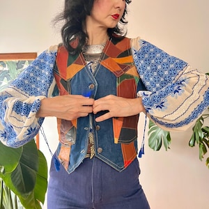 Könnte beinhalten: Eine Patchwork-Weste mit blauen Denim-Einsätzen kombiniert mit braunen, orangefarbenen und schwarzen Lederteilen, die mit Metallknöpfen verschlossen wird. Die Weste wird über einer langärmeligen Bluse aus beigem Stoff mit blauen Mustern getragen.