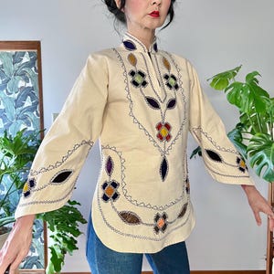 Puede incluir: Túnica color crema con bordados florales multicolores. La parte superior de manga larga presenta un cuello mandarín y costuras decorativas en los bordes. El bordado incluye tonos de azul, amarillo, naranja y morado.