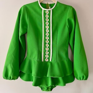 May include: A vibrant green vintage jumpsuit with long sleeves and a ruffled peplum. The neckline and centre front are trimmed with white fabric and daisy-like floral embellishments. The jumpsuit is hanging on a wooden hanger.