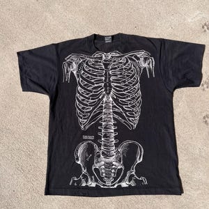May include: Black t-shirt with a white anatomical drawing of a human skeleton. The design shows the rib cage, spine, and pelvis, with anatomical labels. The t-shirt is displayed flat on a textured surface.