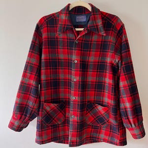 May include: A red, black, and grey plaid button-up shirt with two front pockets. The shirt is hanging on a hanger. The shirt has a collar and long sleeves. The shirt is made of a thick, woven fabric.