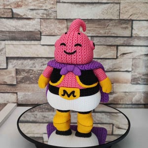 May include: A handmade, pink and purple crochet figure of a character with a smiling face, wearing a black vest, white pants, and yellow boots. The figure has a purple cape and a gold belt with a letter "M".