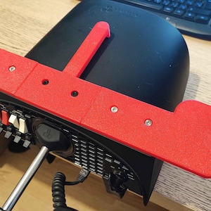 May include: A red 3D-printed bracket with the text "Create3D" is mounted on a black electronic device. The bracket has a rectangular shape with a raised section and several screw holes. The device has buttons and a power cord.