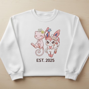 Könnte beinhalten: Weißes Sweatshirt mit einem Cartoon-Design von zwei Charakteren, jeweils mit Blumen geschmückt. Der Text "EST. 2025" ist in Braun gedruckt. Die Charaktere sind rosa und weiß mit großen Augen und in einer spielerischen Pose.