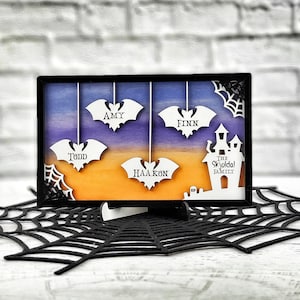 Könnte beinhalten: Ein gerahmtes Halloween-Familienporträt mit Sonnenuntergangs-Hintergrund. Die Namen Amy, Finn, Todd und Haakon sind auf Fledermäusen abgebildet, die von oben hängen. Eine weiße Silhouette eines Hauses mit dem Text "The Skyoldal Family" befindet sich auf der rechten Seite des Rahmens.