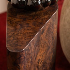 Puede incluir: Una escultura de madera con una rica veta de madera de nogal marrón oscuro. La escultura tiene una parte superior redondeada con una serie de cuentas esféricas oscuras y brillantes. La base tiene forma rectangular con un corte diagonal en un lado.