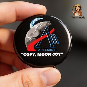 May include: Black round button with the Artemis II logo, featuring a stylized moon and Earth, with red and blue accents. The text "COPY, MOON JOY" is at the bottom. The button is held in a hand.