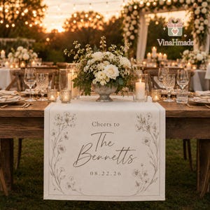 May include: A wedding reception table adorned with a floral arrangement, candles, and glasses. A table runner with floral designs and the text "Cheers to The Bennetts 08.22.26" is visible. The scene is lit with warm, inviting lighting.