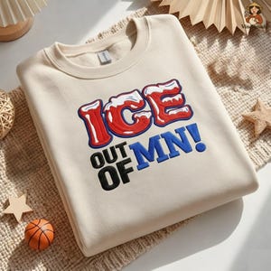 May include: Cream-colored sweatshirt with the text "ICE OUT OF MN!" in red, white, and blue. The sweatshirt has a crew neck and long sleeves. A basketball and star-shaped decorations are in the background.