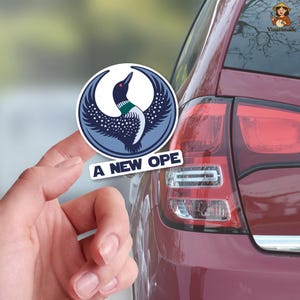 May include: A round sticker featuring a stylized loon with outstretched wings in shades of blue, white, and black. The sticker has the text "A NEW OPE" and is held in front of a red car.