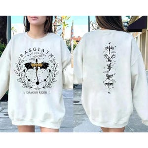 May include: Cream-colored sweatshirt with a graphic design on the front and back. The front features the text "BASGIATH WAR COLLEGE" in a circular design with a dragon and the words "DRAGON RIDER". The back has a vertical dragon design.
