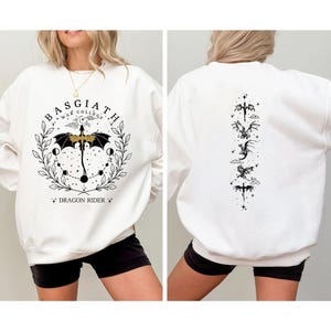 May include: White sweatshirt with a black and gold graphic on the front that says "BASGIATH WAR COLLEGE" with a dragon and umbrella design. The back of the sweatshirt has a vertical design of dragons. The sweatshirt is worn by a person.