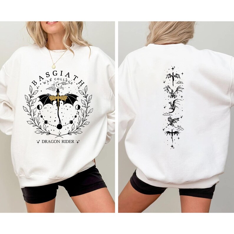 May include: White sweatshirt with a black and gold graphic on the front that says "BASGIATH WAR COLLEGE" with a dragon and umbrella design. The back of the sweatshirt has a vertical design of dragons. The sweatshirt is worn by a person.