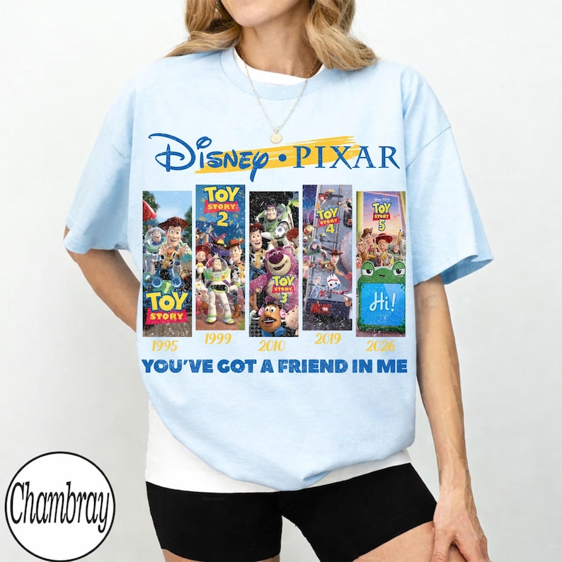 Może przedstawiać: Jasnoniebieska koszulka z logo Disney Pixar i plakatami filmowymi Toy Story. Koszulka przedstawia tytuły film&oacute;w, lata premiery i frazę "YOU'VE GOT A FRIEND IN ME."