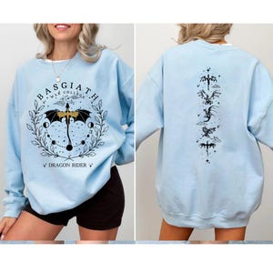 May include: Light blue sweatshirt with a graphic on the front that says "Basgiath War College" with a dragon and umbrella design. The back of the sweatshirt has a vertical design of dragons and other symbols. The sweatshirt is a crew neck.