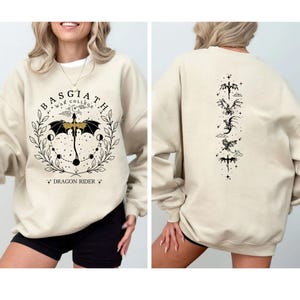 May include: Beige sweatshirt with black graphic design. The front features text that reads "BASGIATH WAR COLLEGE" and "DRAGON RIDER" with a dragon and umbrella design. The back has a vertical arrangement of dragon and sword illustrations.