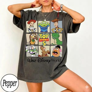 Pode incluir: Camiseta cinza escuro com uma grade de personagens de Toy Story, incluindo Woody, Buzz Lightyear e Rex. A camiseta tem o texto "Toy Story 5" e "Walt Disney World". A camiseta tem um corte folgado.