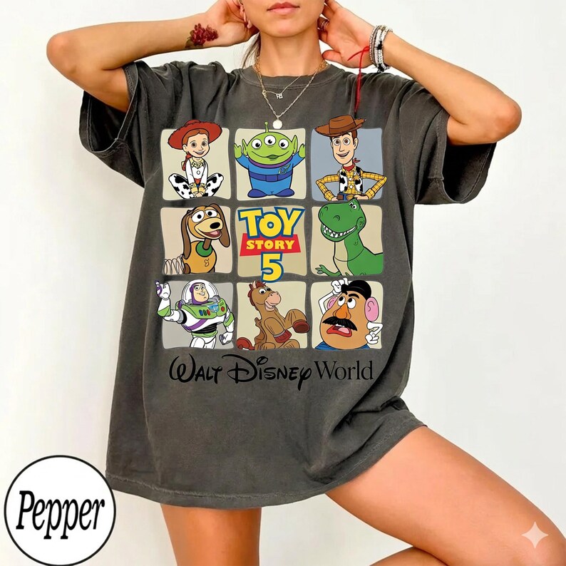 Pode incluir: Camiseta cinza escuro com uma grade de personagens de Toy Story, incluindo Woody, Buzz Lightyear e Rex. A camiseta tem o texto "Toy Story 5" e "Walt Disney World". A camiseta tem um corte folgado.