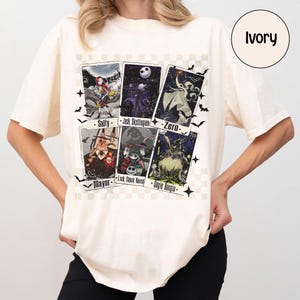 May include: Ivory-colored t-shirt featuring a graphic of six character cards from a movie. The cards include Sally, Jack Skellington, Zero, The Mayor, Lock, Shock, Barrel, and Oogie Boogie. Black bats and stars surround the cards.