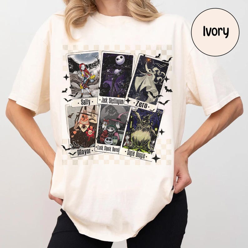 May include: Ivory-colored t-shirt featuring a graphic of six character cards from a movie. The cards include Sally, Jack Skellington, Zero, The Mayor, Lock, Shock, Barrel, and Oogie Boogie. Black bats and stars surround the cards.