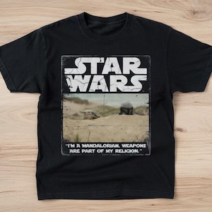 May include: Black Star Wars t-shirt with a rectangular graphic featuring the Star Wars logo and a desert scene. The text "I'M A MANDALORIAN. WEAPONS ARE PART OF MY RELIGION." is printed below the image. The shirt is made of a soft material.
