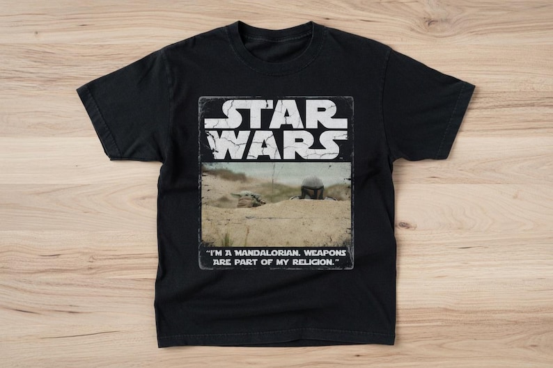 May include: Black Star Wars t-shirt with a rectangular graphic featuring the Star Wars logo and a desert scene. The text "I'M A MANDALORIAN. WEAPONS ARE PART OF MY RELIGION." is printed below the image. The shirt is made of a soft material.