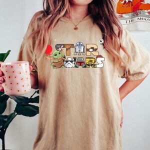 May include: A tan t-shirt with the words "STAR WARS" in a colourful cartoon design. The letters are filled with images of characters including Grogu, R2-D2, and a Stormtrooper. The shirt has short sleeves and a relaxed fit.