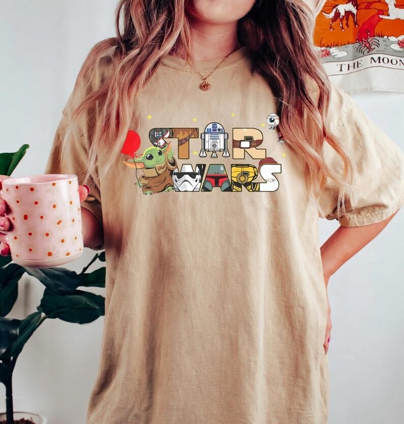 May include: A tan t-shirt with the words "STAR WARS" in a colourful cartoon design. The letters are filled with images of characters including Grogu, R2-D2, and a Stormtrooper. The shirt has short sleeves and a relaxed fit.