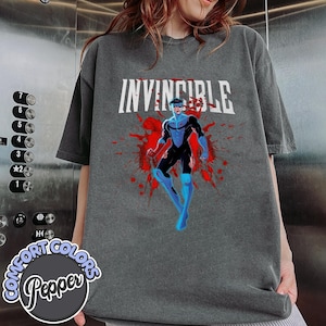 May include: Dark gray t-shirt featuring the word "INVINCIBLE" in white block letters above a graphic of a superhero in a blue and black suit, with red splatter accents. The shirt is a Comfort Colors brand.