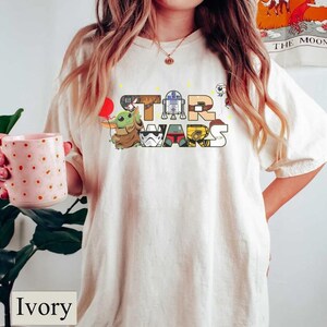 May include: Ivory-coloured t-shirt with the words "STAR WARS" in a colourful, cartoonish font. The letters are decorated with Star Wars characters, including Grogu, R2-D2, and a Stormtrooper. The shirt has a relaxed fit.