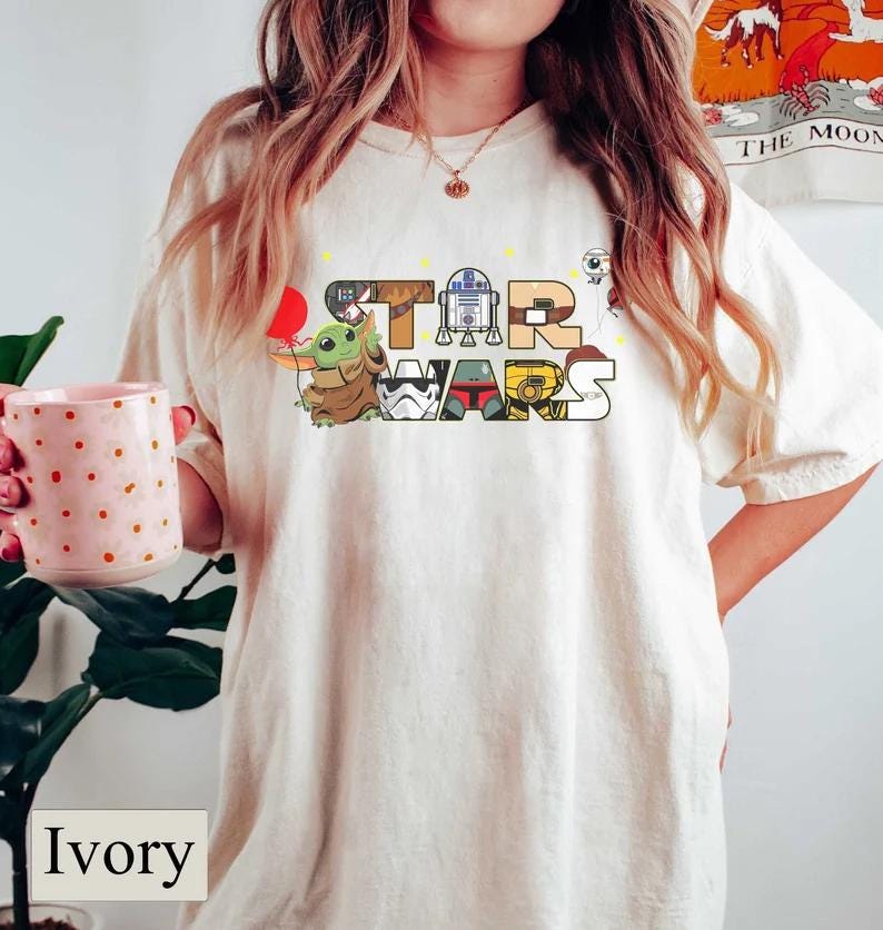 May include: Ivory-coloured t-shirt with the words "STAR WARS" in a colourful, cartoonish font. The letters are decorated with Star Wars characters, including Grogu, R2-D2, and a Stormtrooper. The shirt has a relaxed fit.