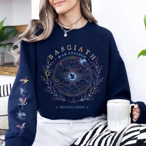 Puede incluir: Sudadera azul marino con un diseño gráfico circular con el texto "BASGIATH WAR COLLEGE" y "DRAGON RIDER". El diseño incluye un motivo de dragón y nubes en colores pastel. La manga izquierda tiene ilustraciones de dragones.