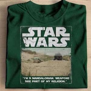 May include: Dark green t-shirt featuring a distressed graphic from the Star Wars franchise. The graphic includes the words "STAR WARS" and an image of a Mandalorian and Grogu. The text "I'M A MANDALORIAN. WEAPONS ARE PART OF MY RELIGION." is also included.