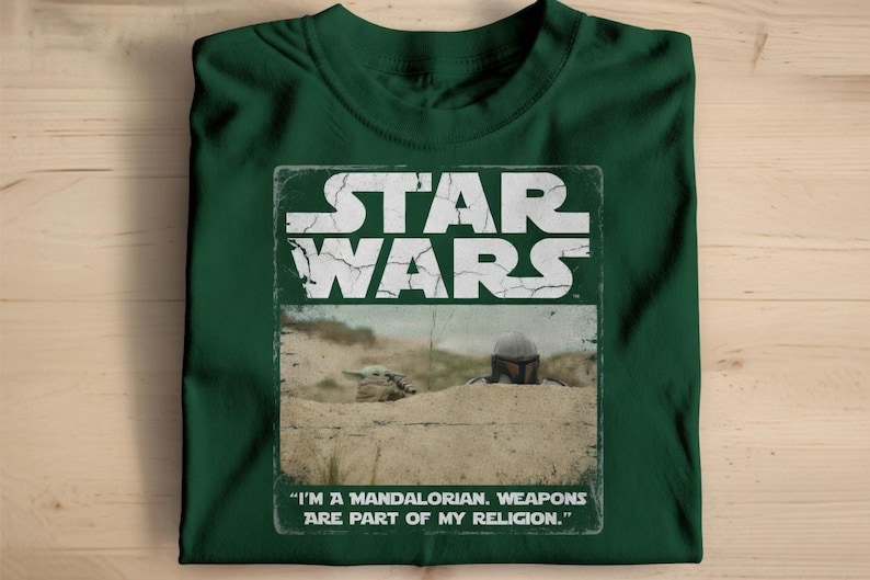 May include: Dark green t-shirt featuring a distressed graphic from the Star Wars franchise. The graphic includes the words "STAR WARS" and an image of a Mandalorian and Grogu. The text "I'M A MANDALORIAN. WEAPONS ARE PART OF MY RELIGION." is also included.