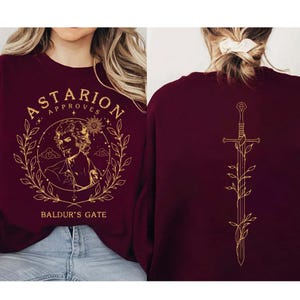 Puede incluir: Sudadera burdeos con ilustraciones y texto dorados. La parte delantera presenta una ilustración de personaje, las palabras "Astarion Approves" y "Baldur's Gate". La parte trasera muestra un diseño de espada dorada con detalles de hojas.