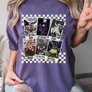 May include: Purple t-shirt featuring playing card designs with characters from The Nightmare Before Christmas. The cards include Sally, Jack Skellington, Zero, Mayor, Lock, Shock, Barrel, and Oogie Boogie. The design is set against a checkered background.