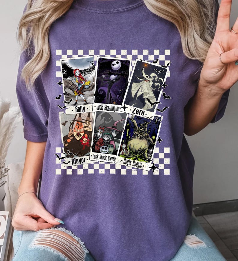 May include: Purple t-shirt featuring playing card designs with characters from The Nightmare Before Christmas. The cards include Sally, Jack Skellington, Zero, Mayor, Lock, Shock, Barrel, and Oogie Boogie. The design is set against a checkered background.