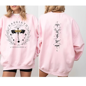 May include: A light pink sweatshirt with a graphic on the front and back. The front features a circular design with the text "BASGIATH WAR COLLEGE" and "DRAGON RIDER". The back has a vertical arrangement of dragon illustrations.