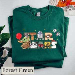 May include: Forest green t-shirt with a colourful "STAR WARS" design featuring cartoon characters. The design includes Baby Yoda with a red balloon, R2-D2, and other characters from the Star Wars franchise.