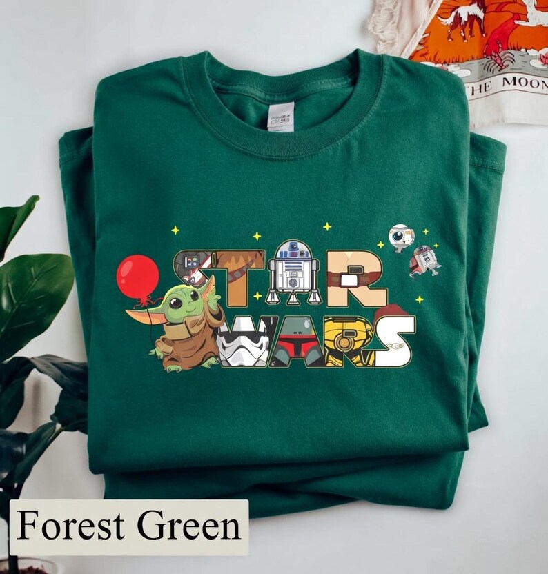 May include: Forest green t-shirt with a colourful "STAR WARS" design featuring cartoon characters. The design includes Baby Yoda with a red balloon, R2-D2, and other characters from the Star Wars franchise.