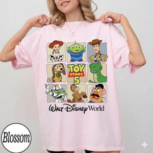 Pode incluir: Camiseta rosa claro com uma grade de personagens de Toy Story, incluindo Woody, Buzz Lightyear e Rex. A camisa tamb&eacute;m tem o texto "Toy Story 5" e "Walt Disney World" impresso.